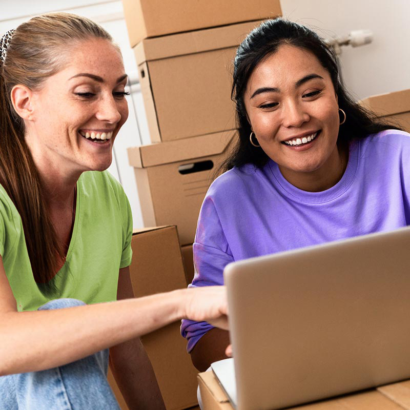 Friends set up WiFi on a laptop in a new apartment surrounded by moving boxes